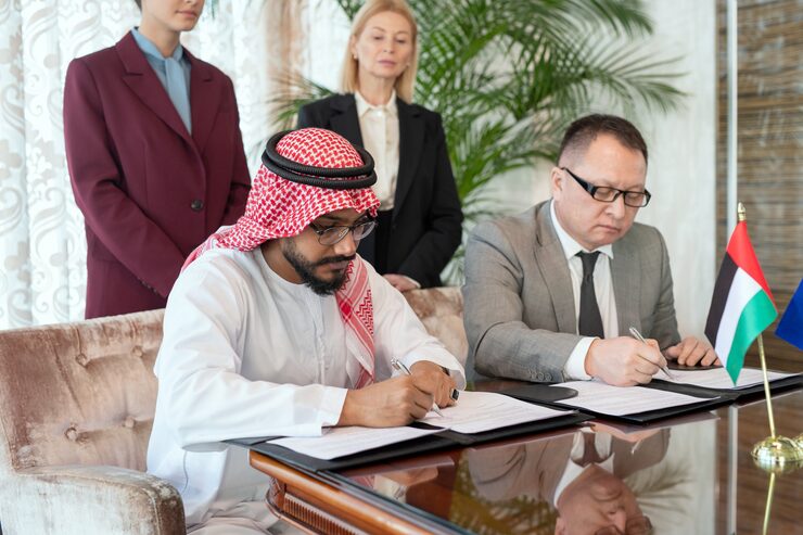 two-serious-business-partners-sitting-by-table-front-camera-signing-contract-with-elegant-businesswomen-standing-background_274679-18344